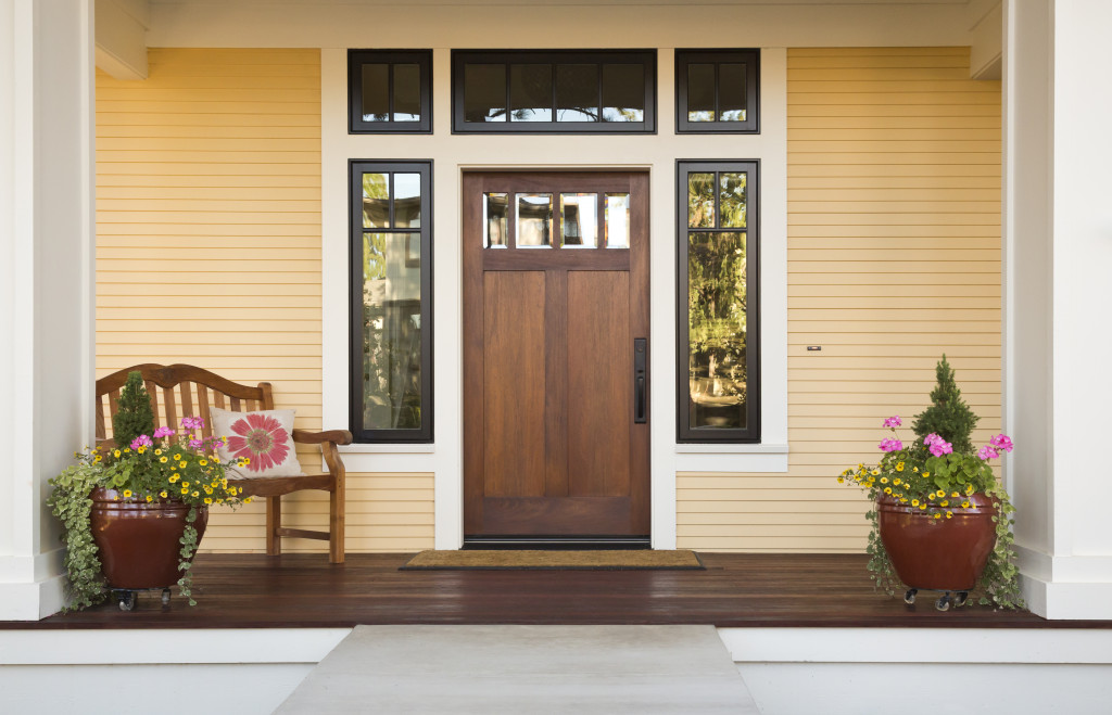 Front view of a wooden front door on a yellow house with reflections in the window and a wide view of the porch and front walkway.