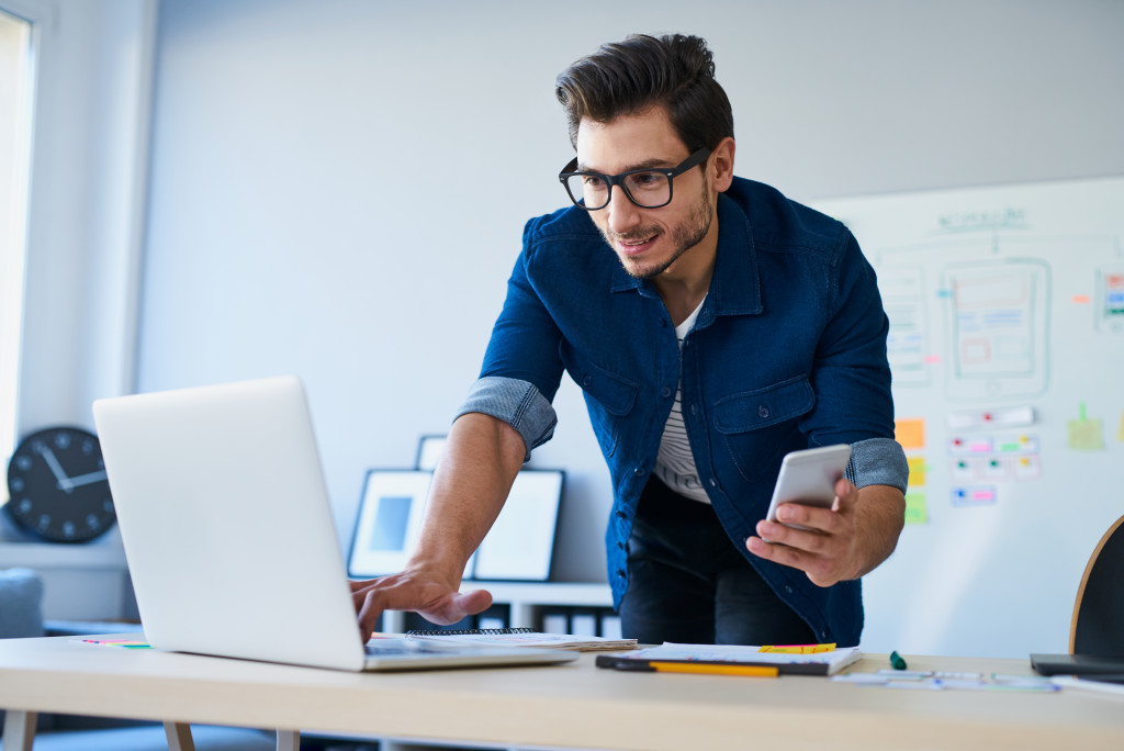 Man checking his laptop while holding a smartphone.